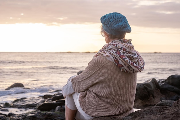 Vue arrière d'une femme senior caucasienne détendue assise sur la plage de galets à la lumière du coucher du soleil admirant l'horizon au-dessus de l'eau Dame âgée souriante appréciant la nature et la liberté