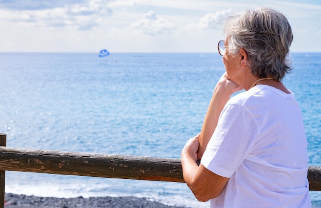 Vue arrière d'une femme senior caucasienne vêtue de blanc détendu appuyé contre un balcon en bois face à la mer regardant l'horizon au-dessus de l'eau