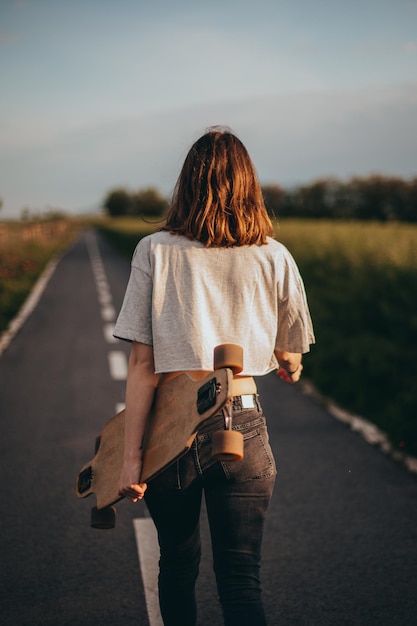 Vue arrière femme skateur tenir longboard sur la piste cyclable pré vert en arrière-plan haut q