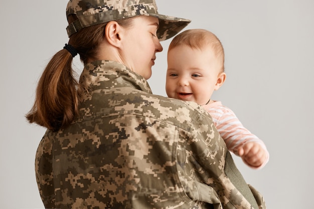 Vue arrière d'une femme soldat portant un uniforme de camouflage et une casquette, serrant sa petite fille dans ses bras tout en la tenant dans les mains, rentrant chez elle de l'armée ou de la guerre.