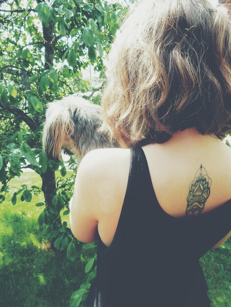 Vue arrière d'une femme avec un tatouage sur le dos tenant un chien contre un arbre