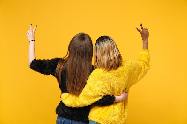 Vue arrière femmes amis européens et afro-américains en vêtements jaunes noirs debout posant isolés sur fond de mur orange vif, portrait en studio. Concept de style de vie des gens. Maquette de l'espace de copie.