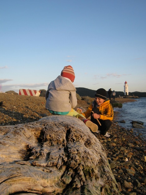 Photo vue arrière d'une fille assise sur du bois flottant par un frère souriant jouant à la plage