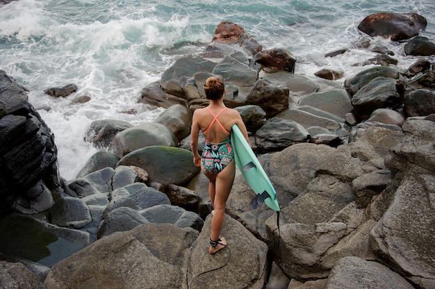 Vue arrière fille en maillot de bain multicolore marchant avec le surf sur la plage rocheuse de l'océan Atlantique en regardant au loin