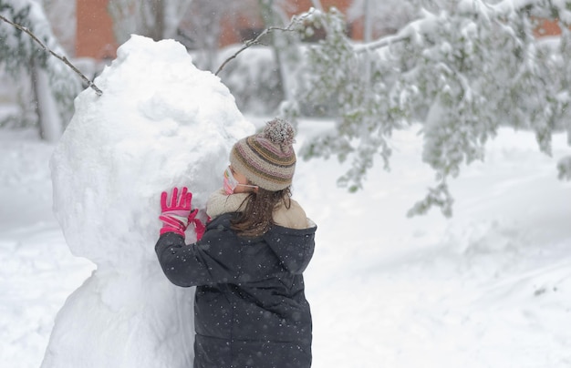 Vue arrière d'une fille portant un masque de coronavirus construisant un bonhomme de neige lors d'une chute de neige.