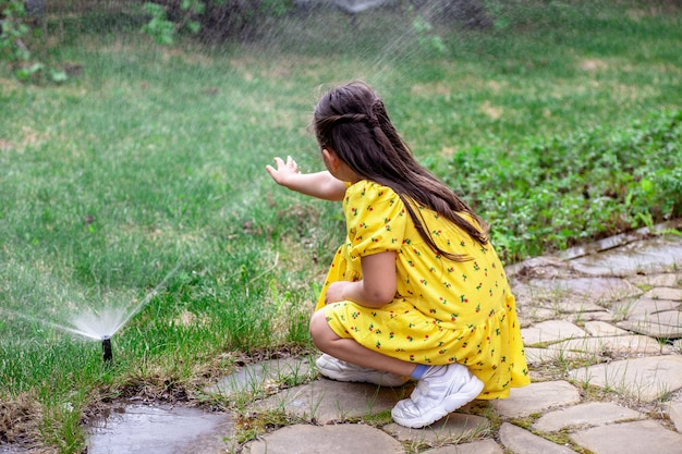 Vue arrière d'une fille qui joue avec les gouttes d'eau d'un arroseur de pelouse une fille aide ses pa...