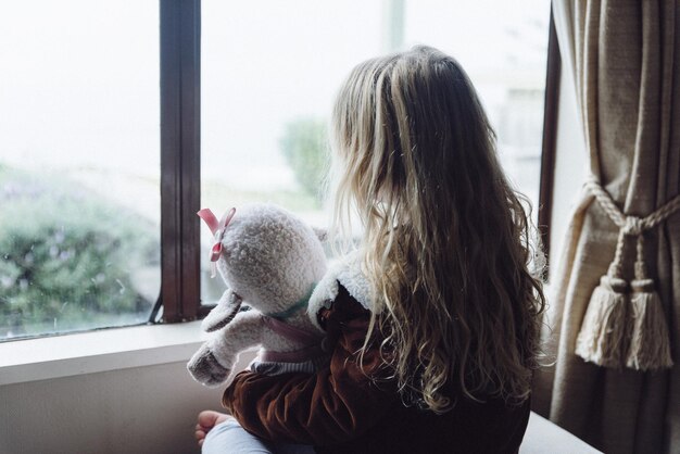 Vue arrière d'une fille tenant un jouet en peluche en regardant par la fenêtre