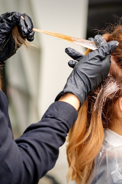 Vue arrière en gros plan des mains de coiffeurs dans des gants appliquant un colorant sur une mèche de cheveux d'une jeune femme rousse dans un salon de coiffure