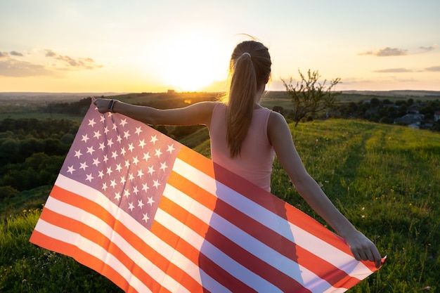 Vue arrière de l'heureuse jeune femme posant avec le drapeau national des USA debout à l'extérieur au coucher du soleil.