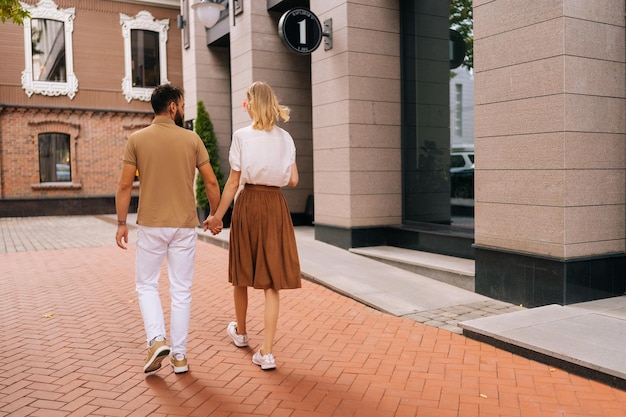 Vue arrière de l'heureux jeune couple amoureux portant des vêtements décontractés marchant ensemble dans la rue de la ville, main dans la main