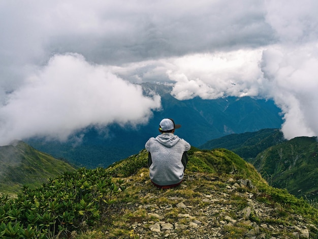 Vue arrière de l'homme actif assis sur le sommet de la montagne et se détendre au fond des nuages