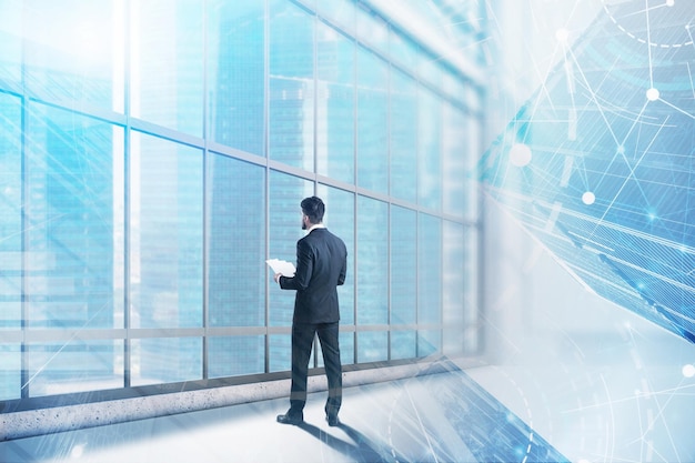 Vue arrière d'un homme d'affaires avec des documents debout dans un bureau panoramique regardant la ville avec une double exposition d'une interface immersive bleue. Concept de haute technologie. Image tonique