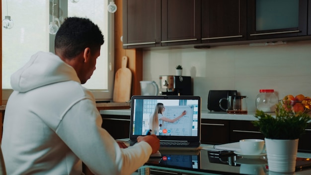 Vue arrière d'un homme africain regardant une vidéoconférence en ligne sur ordinateur et prenant des notes. Il travaille à la maison en quarantaine.