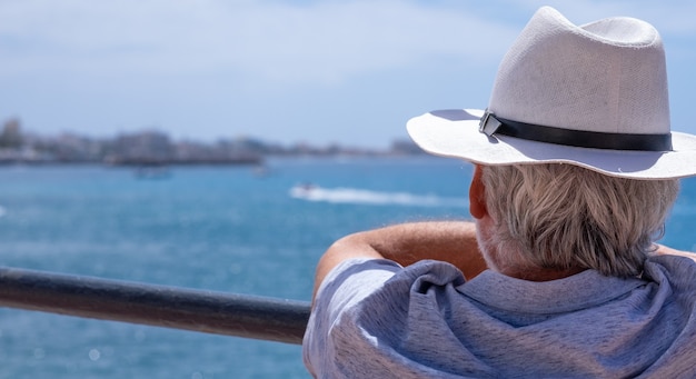 Photo vue arrière de l'homme aux cheveux gris avec un chapeau regardant le paysage marin et l'horizon au-dessus de l'eau en vacances à la mer