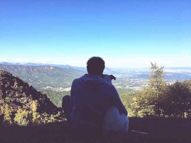 Vue arrière d'un homme avec un chien regardant la vue alors qu'il est assis sur la montagne contre un ciel clair