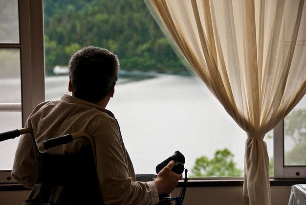 Photo vue arrière d'un homme en fauteuil roulant regardant la rivière par la fenêtre de sa maison