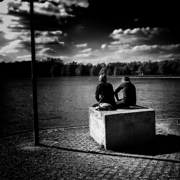 Photo vue arrière d'un homme et d'une femme assis au bord d'un lac contre le ciel