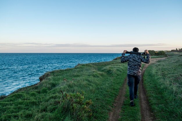 Photo vue arrière d'un homme portant un trépied sur un sentier par la mer contre le ciel