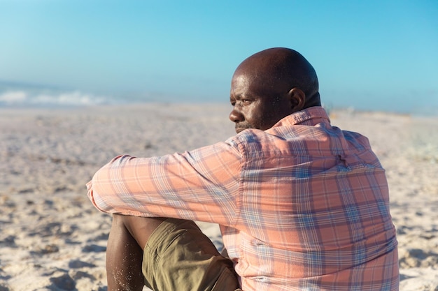 Vue arrière d'un homme senior afro-américain à la retraite regardant ailleurs assis sur la plage par une journée ensoleillée