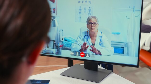 Vue arrière d'une infirmière discutant sur webcam avec un stomatologue spécialiste senior utilisant un téléphone portable assis dans une clinique dentaire devant un ordinateur avec radiographie numérique. Médecin dentiste expliquant les symptômes du patient