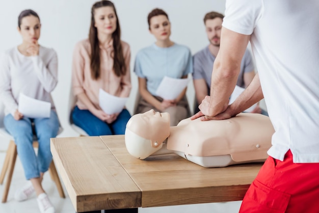 Vue arrière de l'instructeur exécutant la RCR sur un mannequin pendant la formation aux premiers secours avec un groupe de personnes