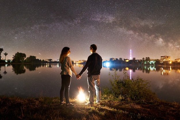 Vue arrière de la jeune famille - homme et femme debout sur une rive du lac près d'un feu de joie, se tenant la main, profitant d'une vue magnifique sur le ciel nocturne plein d'étoiles et la voie lactée au-dessus de l'eau calme et de la ville lumineuse