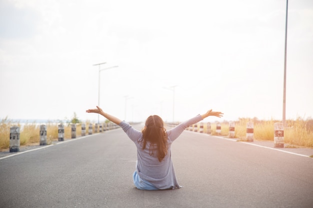 La vue arrière d'une jeune femme assise sur la route, regardant loin et les mains vers le haut.