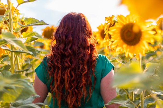 Vue arrière d'une jeune femme aux cheveux bouclés rouges et une robe verte posant dans un champ de tournesols