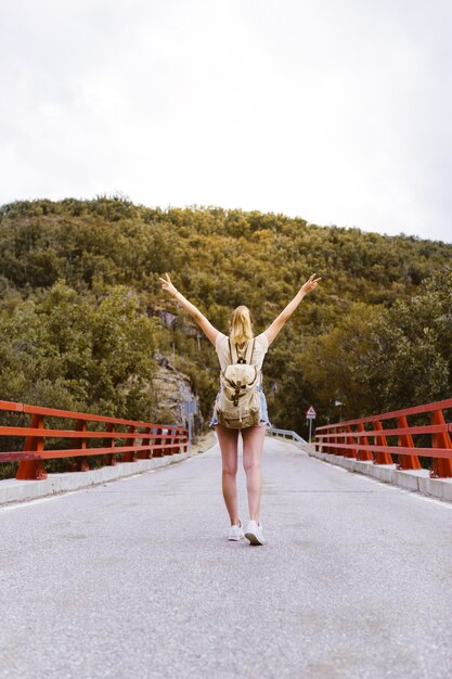 Vue arrière de la jeune femme blonde avec sac à dos et mains en marchant sur la route sur un pont près de la montagne. Concept de voyage et d'aventure. Voyageur au milieu des bois. Voyager seul