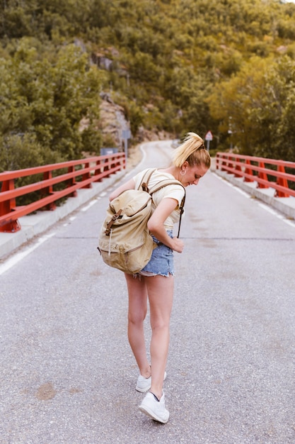 Vue arrière de la jeune femme blonde avec sac à dos marchant sur la route sur un pont près de la montagne. Concept de voyage et d'aventure. Voyageur au milieu des bois. Voyager seul