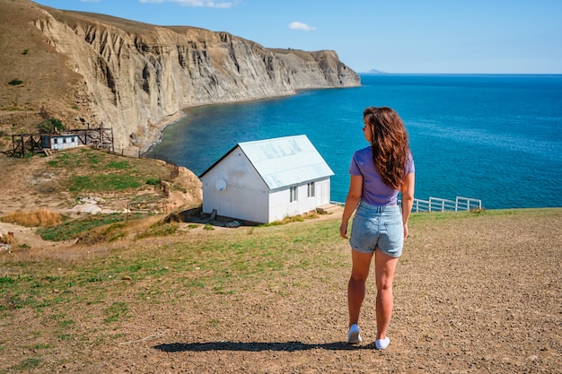 Vue arrière d'une jeune femme devant une maison solitaire blanche au bord d'une falaise