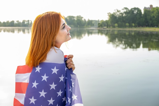 Vue arrière d'une jeune femme heureuse avec le drapeau national des États-Unis sur ses épaules. Fille positive célébrant le jour de l'indépendance des États-Unis.