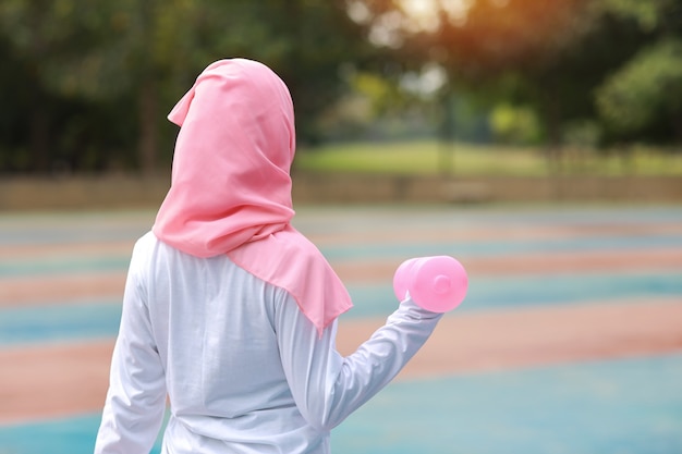 Vue arrière jeune femme musulmane asiatique en sportswear debout et soulevant des haltères en plein air pour l'exercice du matin.