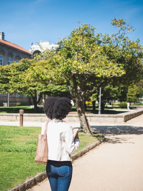 Vue arrière d'une jeune femme noire, aux cheveux bouclés, tenant des livres, à l'extérieur