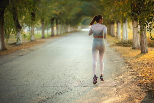 Vue arrière d'une jeune femme portant des vêtements de sport fait du jogging le long du sentier de l'avenue ensoleillée.