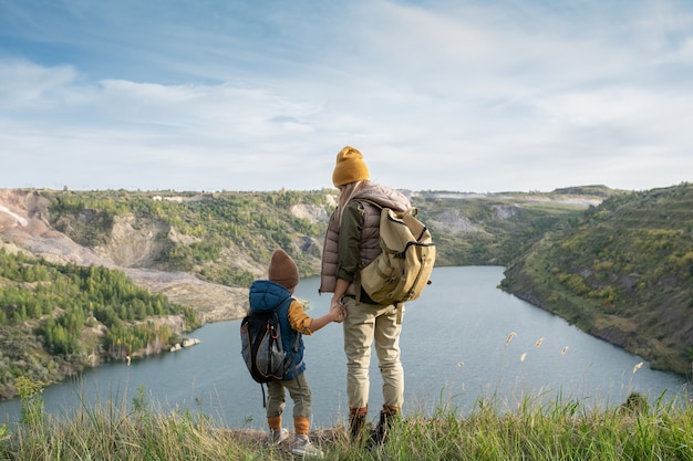 Vue arrière d'une jeune femme routard et d'un petit garçon tenant par les mains tout en se tenant devant le lac entouré de montagnes pendant le voyage