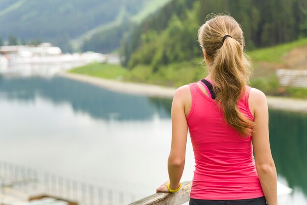 Vue arrière d'une jeune femme sportive dans des vêtements de fitness debout à l'extérieur près du grand lac dans les montagnes, profitant de la vue sur la nature.