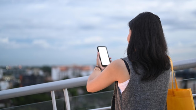 Vue arrière d'une jeune femme utilisant un téléphone portable debout sur une terrasse avec un beau ciel du soir en arrière-plan