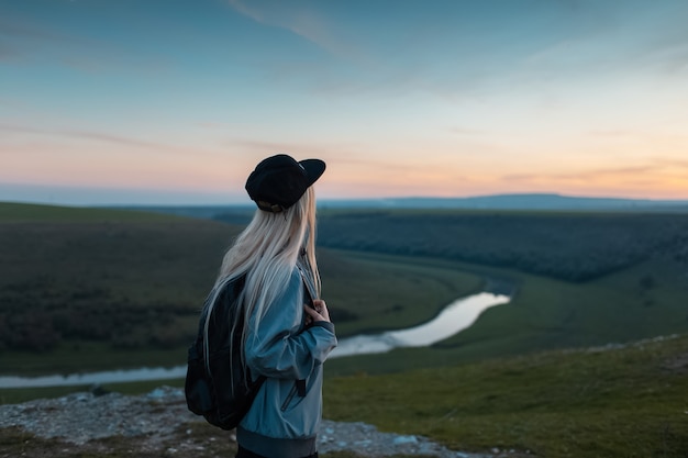 Vue arrière de la jeune fille blonde avec sac à dos, profitant du coucher du soleil sur le sommet des collines. Concept de voyage.