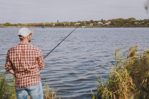 Vue arrière Jeune homme mal rasé avec canne à pêche en chemise à carreaux, casquette et lunettes de soleil jette des appâts et pêche sur le lac depuis la rive près des arbustes et des roseaux. Mode de vie, loisirs, concept de loisirs de pêcheur.