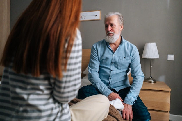 Vue arrière d'une jeune petite-fille aimante parlant avec un grand-père aux cheveux gris assis dans son lit dans une chambre confortable