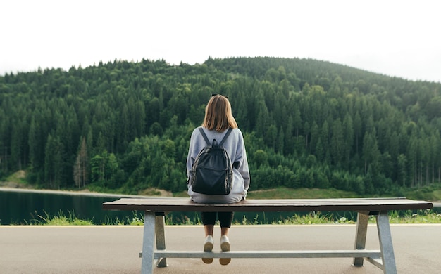 Vue arrière d'une jeune voyageuse assise seule sur le banc en profitant de la magnifique vue sur la forêt de sapins