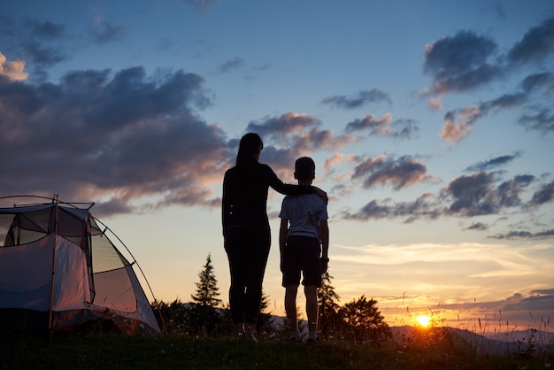 Vue arrière de la mère embrasse son fils près de la tente au sommet de la montagne en profitant de la vue du coucher du soleil, en arrière-plan - montagnes et collines sous un ciel du soir incroyablement beau avec des nuages