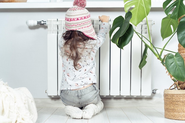 Vue arrière petite fille bouclée congelée en bonnet tricoté chaud s'asseoir sur les genoux mains chaudes sur le radiateur de chaleur Économie de gaz et de chauffage