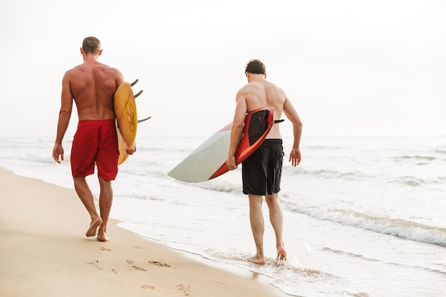 Vue arrière photo d'un jeune deux hommes amis surfeurs avec des surfs sur une plage à l'extérieur.