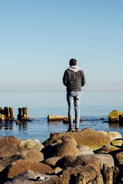 Vue arrière en pleine longueur de l'homme debout sur des rochers par la mer contre un ciel clair