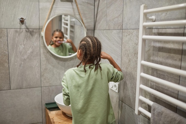 Vue arrière portrait d'enfant noir avec des cheveux tressés se brosser les dents par miroir dans l'espace de copie de la salle de bain