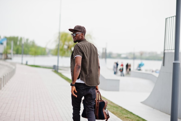 Vue arrière portrait d'un homme afro-américain élégant portant des lunettes de soleil et une casquette avec sac à main en plein air Street fashion black man