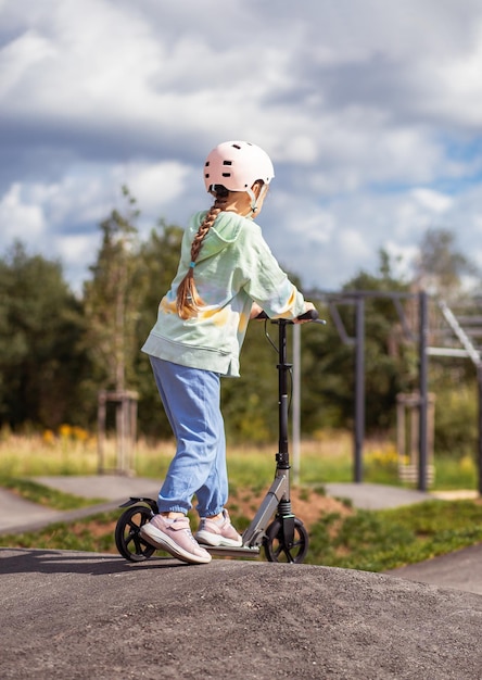 Vue arrière portrait d'une jolie petite écolière caucasienne porter un casque s'amuser à faire du scooter sur le parc de rue asphalté trackxAin à l'extérieur le jour ensoleillé activités sportives saines pour les enfants à l'extérieur
