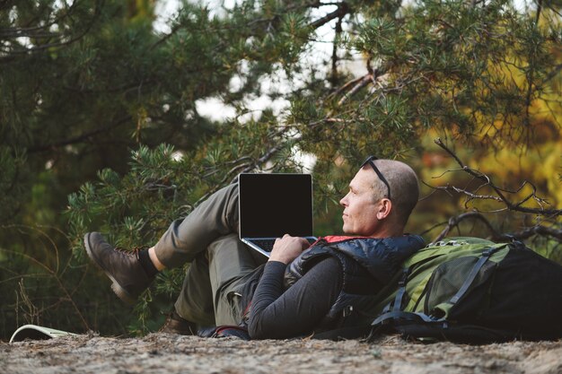 Vue arrière, randonneur masculin allongé sur le dos appuyé sur un sac à dos, avec ordinateur portable, dactylographie, blogs, navigation en forêt